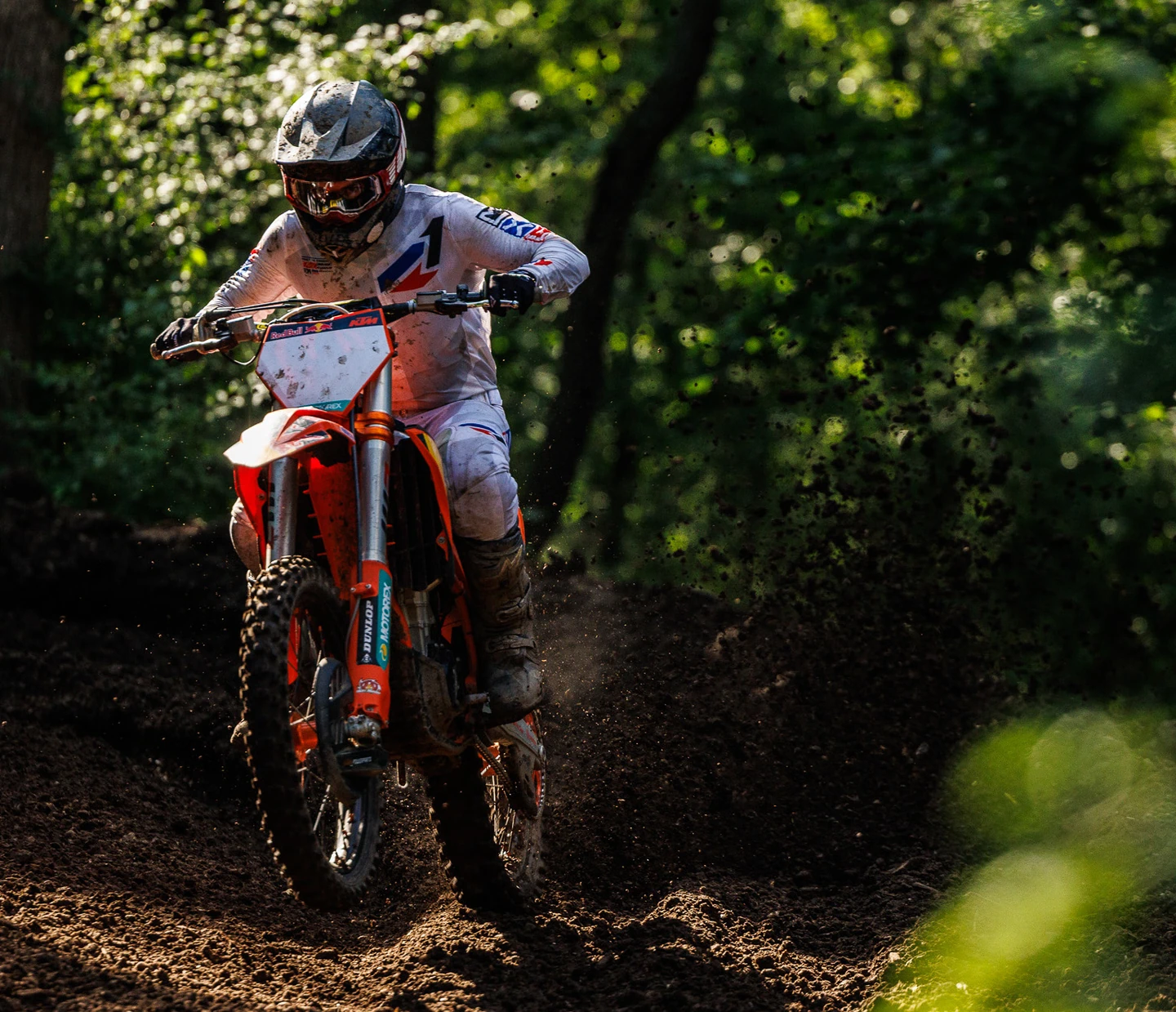 Motocross rider on orange dirt bike kicking up mud in forest.