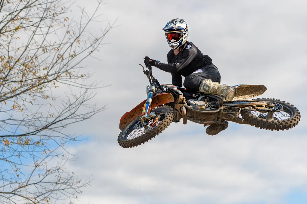 Motocross rider mid-air on muddy dirt bike against cloudy sky.