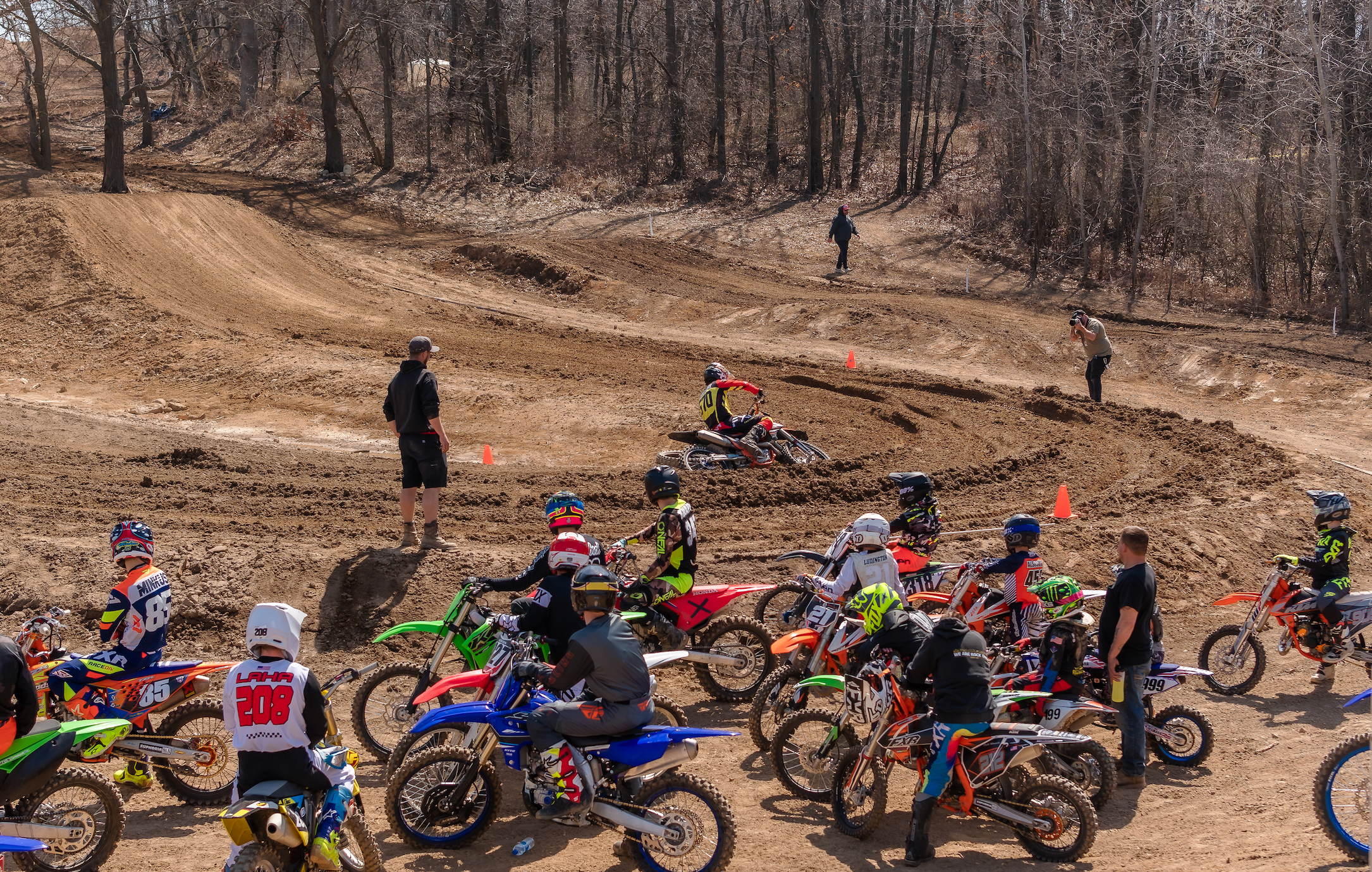 Motocross racers and spectators on a dirt track with bare trees.